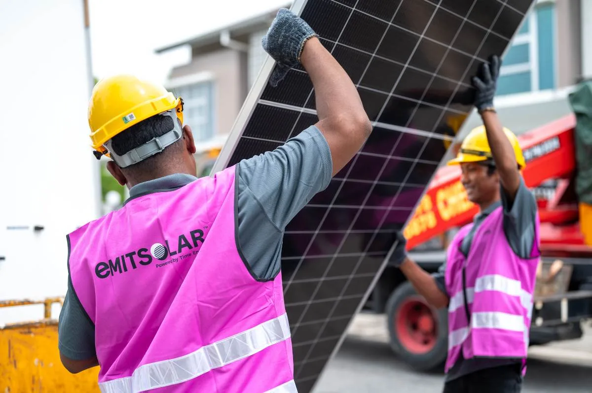 Two Emit Solar installers in pink vests carrying a solar panel to a home
